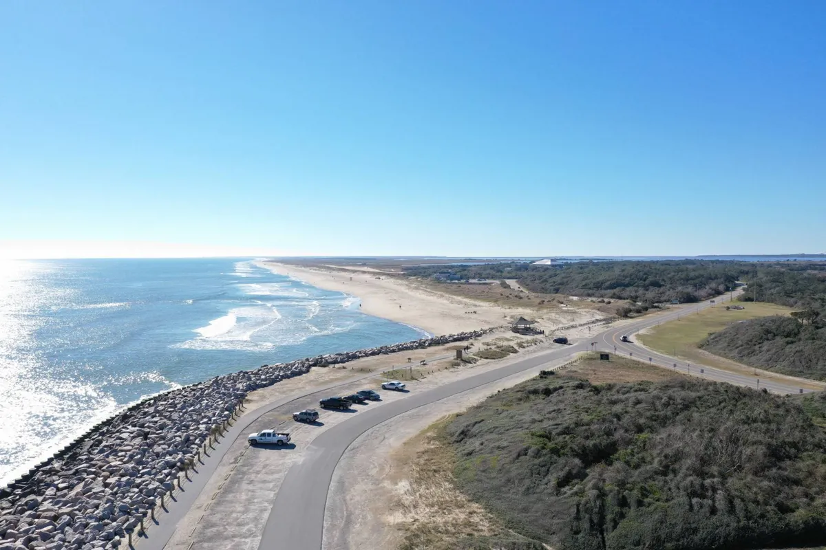 A dynamic scene featuring ocean waves, a sandy shoreline, vehicles parked along the road, and jetty rocks—indicative of an active beach environment. People can be seen walking along the beach and enjoying various water sports, illustrating the adventurous side of the location.