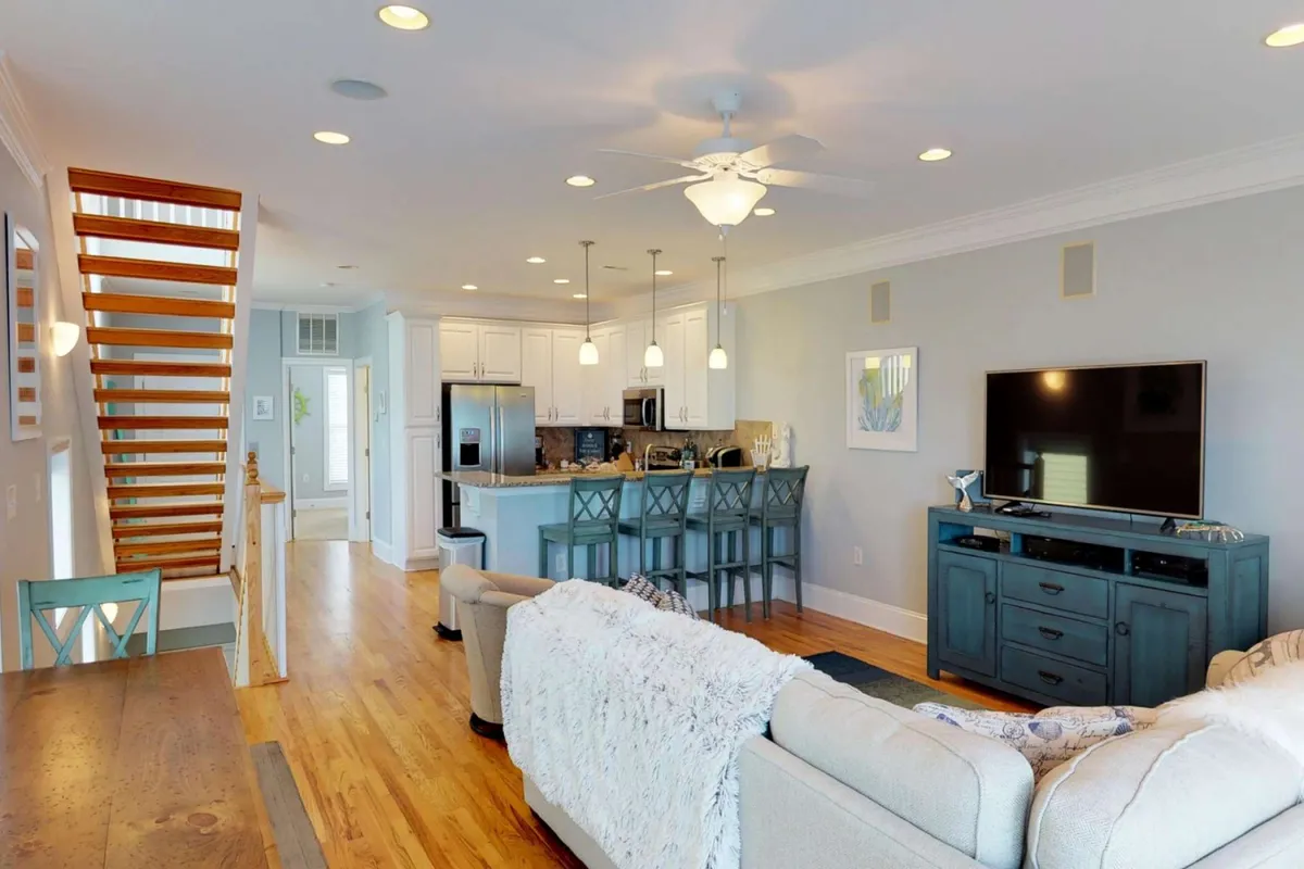 This image captures an open-plan interior with a comfortable sofa, kitchen island, dining table, a visible staircase, and a large television. The arrangement suggests a seamless flow between living, dining, and culinary spaces, bathed in natural light, with space for family gatherings or entertaining friends.