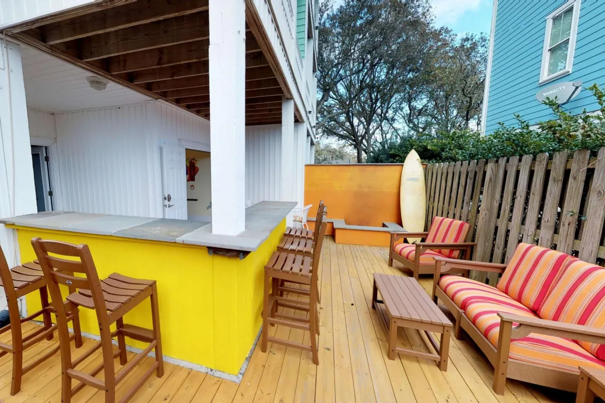 This image features a bright yellow tiki bar counter with striped lounge chairs and a surfboard propped against a fence. The space is set outdoors, accented by sunny skies and palm fronds, with an inviting, casual vibe perfect for gatherings.