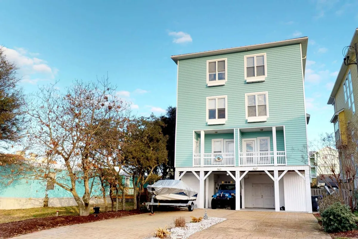 A cheerful blue house with a welcoming porch, a boat in the driveway, and ample yard space. The scene captures the spirit of relaxed gatherings, outdoor activities, and sunset views from the porch.