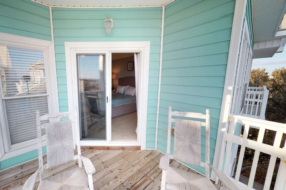 A tranquil porch scene with turquoise walls, a pair of classic white rocking chairs, and a sliding glass door opening to refreshing sea breezes. The porch is shaded and oriented for quiet relaxation or socializing, with subtle coastal accents.