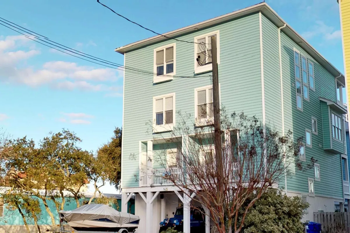 A light blue house with white trim and a boat parked underneath, surrounded by open space and a porch offering views of the outdoors. The inviting exterior suggests a welcoming home base for gatherings, BBQs, and relaxing after a day at the beach.