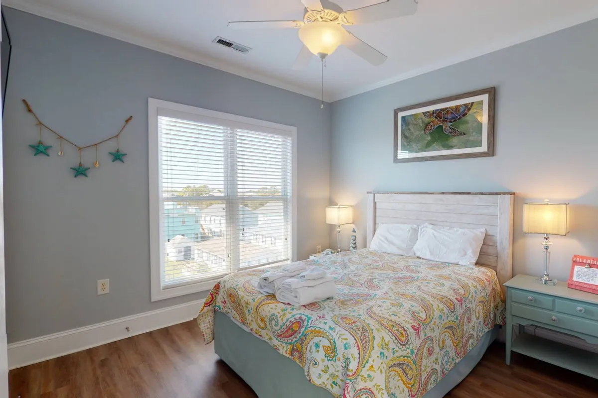 A comfortable bedroom with colorful bedding, framed turtle artwork on the wall, matching side tables with lamps, and natural light streaming through a window. The space feels peaceful and well-appointed for restful nights.