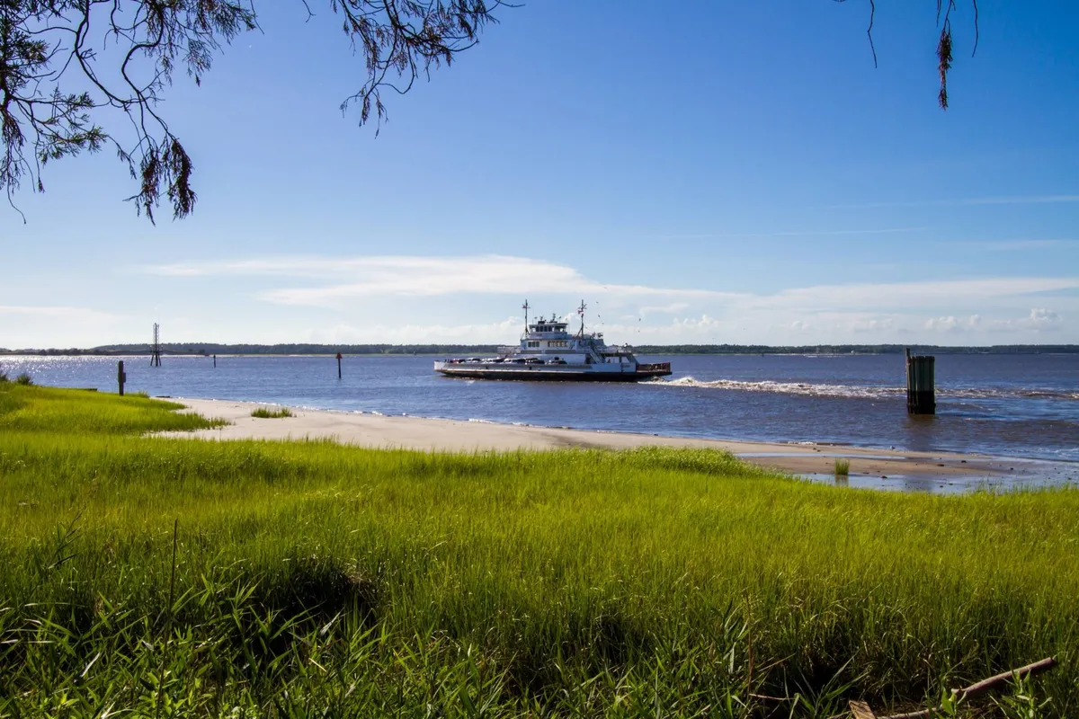 A picturesque view of a ferry gliding across calm water, framed by lush green grass and a clear blue sky. The tranquil setting showcases the local ferry experience and highlights the scenic, low-key outings available nearby.