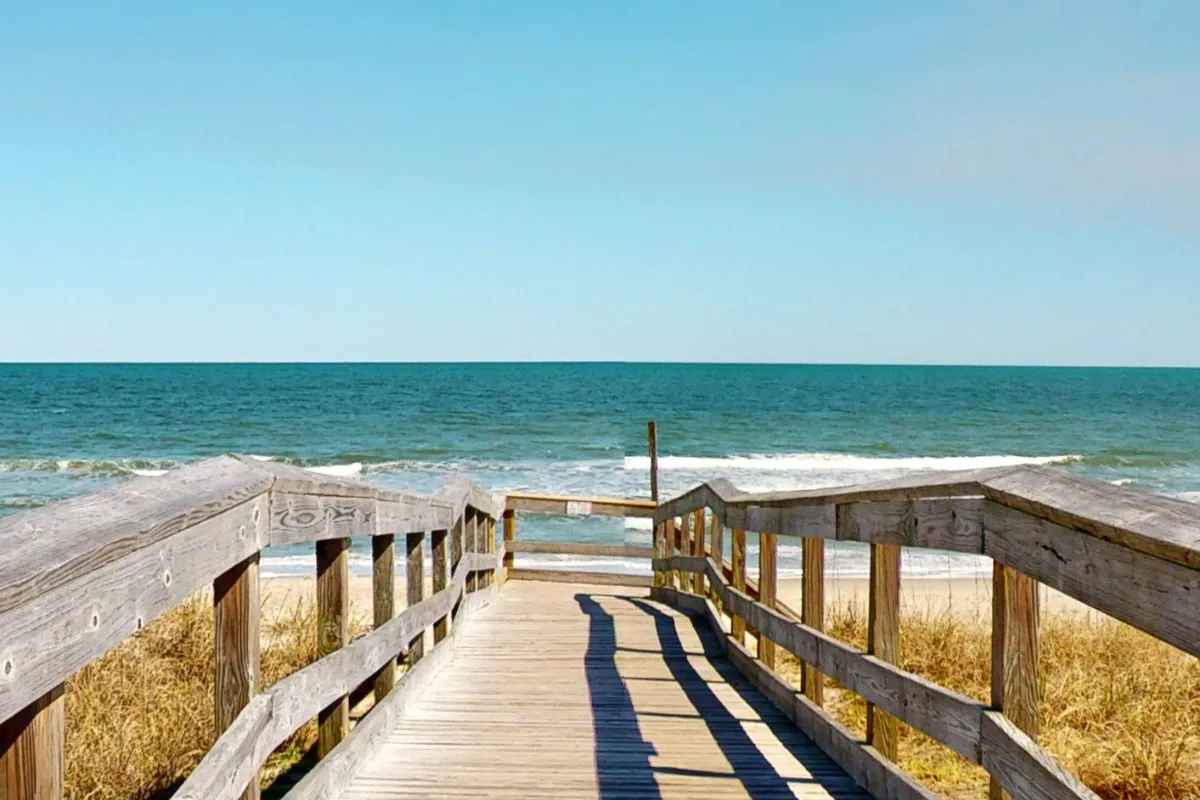 This image features a wooden walkway leading over sand dunes toward the ocean, with the sandy beach and gentle waves visible ahead. The tranquil setting suggests easy, direct access to the beach and evokes the anticipation of family outings, sandcastle building, and seashell collecting.