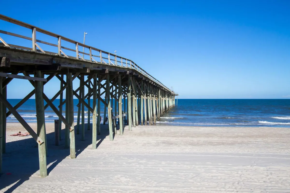 A tranquil pier stretching into the ocean, with soft sand and the gentle waves creating a serene backdrop. The pier is ideal for walks, photography, or simply watching the sunset.