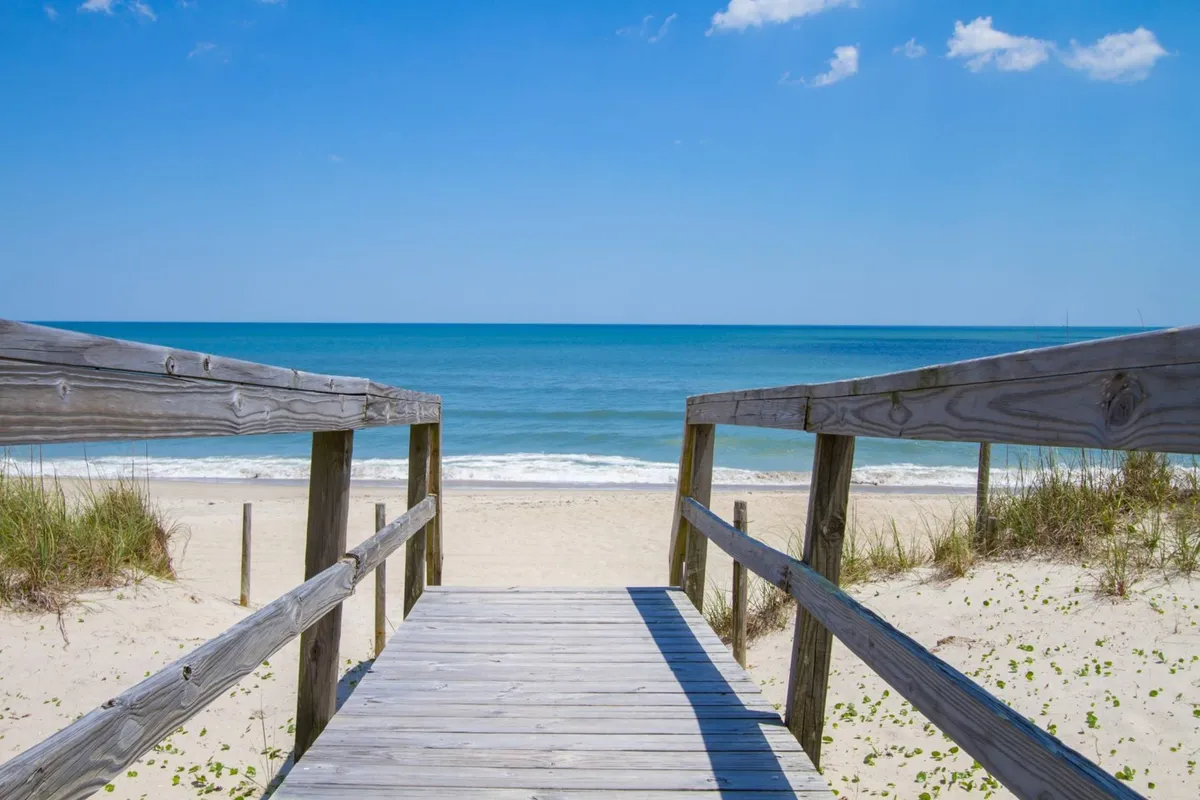 A tranquil wooden walkway leads over dune grasses towards a sandy beach and shimmering ocean. The scene radiates calm, with no crowds present, and offers a gentle, accessible entry to the shore. Natural elements and the inviting pathway highlight the ease of outdoor exploration.
