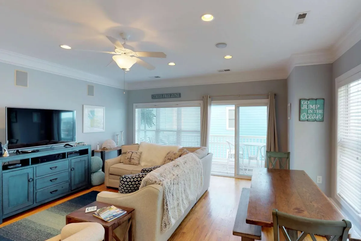 A bright, calm interior showing a plush sofa, large dining table, wall-mounted TV, and sliding glass doors that bring in natural light. The arrangement fosters togetherness and is perfect for watching movies, sharing meals, or simply relaxing as a group.