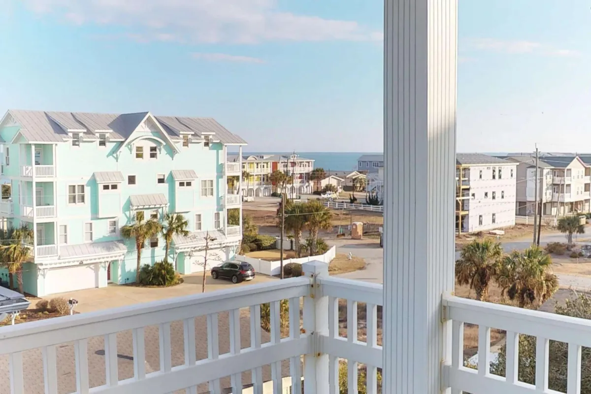 A scenic view of colorful coastal houses with palm trees, all facing a wide expanse of the ocean. The shot is taken from a balcony, emphasizing the peaceful seaside vibe and the opportunity for guests to relax together while overlooking the water.